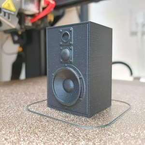 May include: A black speaker with a single large speaker and two smaller speakers. The speaker is sitting on a brown surface.