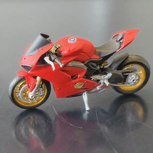 Peut inclure: Une moto jouet rouge et noire avec des roues dorées. La mini moto sportive a un design élégant, avec un pare-brise et une selle noirs. Le modèle est une réplique détaillée d'une moto de course.