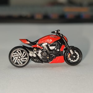 Peut inclure: Modèle réduit de moto Ducati rouge et noire. Le modèle présente des composants détaillés, notamment le moteur, les roues et les tuyaux d'échappement. Le mot "DUCATI" est visible sur le réservoir. La moto a un design élégant et moderne.