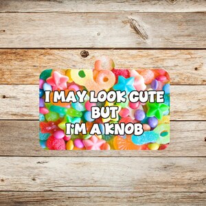 May include: A rectangular sticker featuring a vibrant mix of sweets, such as gummy rings, star-shaped candies, and jelly beans. The sticker displays the text "I MAY LOOK CUTE BUT I'M A KNOB" in white.