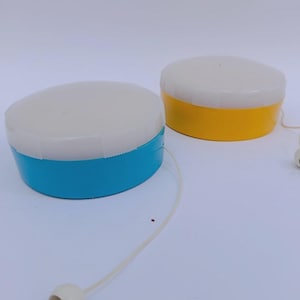 May include: Two vintage yo-yos. One is blue, the other is yellow. Both have a white top and a string with a white button at the end. The yo-yos are on a white background.