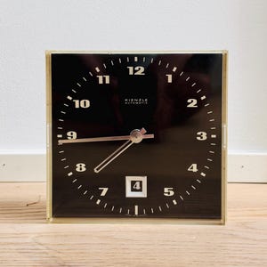 May include: A square Kienzle Automatic desk clock with a black face, white Arabic numerals, and silver-toned hands. The clock features a date window at the six o'clock position and is encased in a clear, transparent frame with a minimalist design.