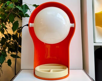 Könnte beinhalten: Eine Tischleuchte mit einem leuchtend orangefarbenen U-förmigen Rahmen, der einen kugelförmigen weißen Glasschirm hält. Der Sockel der Lampe ist eine cremefarbene, kreisförmige Ablage mit einem unterteilten Innenbereich. Ein schwarzes Kabel ist sichtbar.