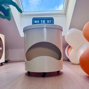 May include: A cylindrical off-white storage unit with a large semi-circular cutout on the front. The unit features a flat top, a smooth finish, and is supported by small black wheels at the base. A blue sign with the text MER 16 SET is visible behind.