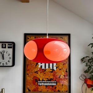 May include: A red, retro-style pendant light with two orange-tinted, oval-shaped glass globes. The light hangs from a white cord. A framed poster with the text "The Virgin Suicides" is visible in the background.