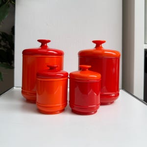 Puede incluir: Cuatro botes de cerámica rojos y naranjas con tapas. Los botes son cilíndricos con base redondeada y un pequeño asa en la tapa. Los colores van del rojo intenso al naranja brillante.