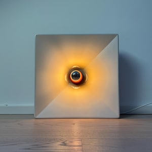 Può includere: Una lampada da parete quadrata bianca con una sorgente luminosa circolare centrale che emette un caldo bagliore arancione. L'apparecchio presenta un design geometrico concavo che irradia la luce verso l'esterno dal centro.