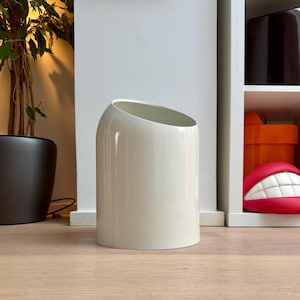 Puede incluir: Un cubo de basura cilíndrico con un acabado liso en color blanco roto. El recipiente presenta una abertura superior inclinada distintiva, creando un perfil asimétrico. La estructura es sólida y opaca, con una superficie exterior curva y limpia.