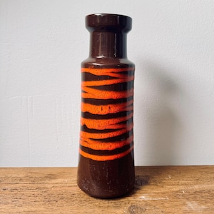 Pode incluir: Um vaso de cerâmica marrom com listras laranja. O vaso tem um fundo arredondado e uma parte superior ligeiramente alargada.