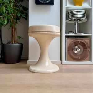 Pode incluir: Um banco creme de estilo moderno de meados do século, com um tampo arredondado e uma base alargada. O banco tem aproximadamente 46 cm de altura. Um relógio quadrado preto com marcações brancas está pendurado na parede atrás do banco.