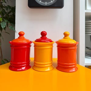 Peut inclure: Trois bocaux en céramique colorés en rouge, jaune et orange avec des couvercles assortis. Une horloge carrée noire avec des marques et des aiguilles blanches est accrochée au mur au-dessus des bocaux. Les bocaux sont posés sur une surface jaune vif.