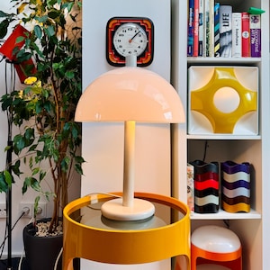 Puede incluir: Una lámpara de mesa blanca con forma de seta, con una pantalla en forma de cúpula y una base cilíndrica, situada sobre una mesa auxiliar circular de color amarillo brillante. La lámpara emite una luz suave y cálida, iluminando el entorno.