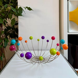 May include: A modern, silver-toned metal fruit bowl with colorful, spherical accents. The bowl features a radial design with various sized spheres in shades of pink, purple, green, orange, and blue. The bowl is on a white surface.