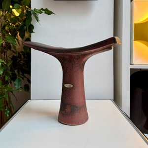 May include: A maroon ceramic vase with a flared top and a narrow base. The vase has a textured surface and a label near the base. A black square clock with white markings hangs on the wall in the background.