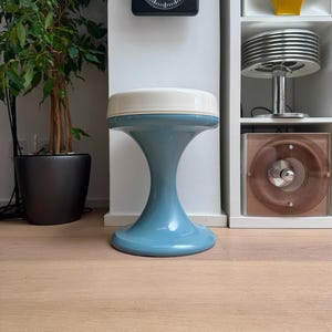 Op de afbeelding: Een lichtblauwe en witte kruk met een unieke zandloper vorm. De kruk heeft een ronde basis en een cirkelvormige bovenkant. Een zwarte vierkante klok met een witte wijzerplaat hangt aan de muur achter de kruk.