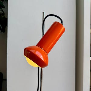 May include: An orange desk lamp with a cylindrical head and a curved, adjustable arm. The lamp has a bright, focused light and a black power cord. The base is a silver pole with a clamp. The lamp is a retro design.