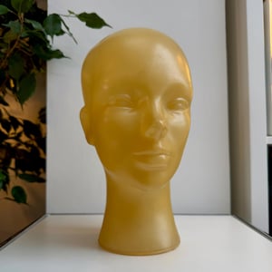 May include: A golden-yellow mannequin head, featuring a smooth, bald surface and facial features. The head is positioned on a white surface, with a green plant visible in the background. This item is likely used for displaying wigs, hats, or other headwear.
