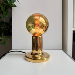 May include: A gold-colored table lamp with a round, globe-shaped bulb. The bulb is lit, emitting a warm glow. The lamp has a cylindrical base and a circular foot. The lamp is on a white surface.