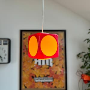 May include: A red cube-shaped pendant light fixture with two orange circular accents on each visible side. The light hangs from a white cord. The fixture is suspended in front of a framed poster.