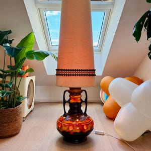 May include: A tall floor lamp featuring a ceramic base with a dark brown and vibrant orange glaze, accented by two handles. The lamp is topped with a tall, cylindrical beige fabric lampshade that is decorated with a dark brown trim band.