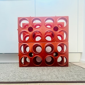 May include: Two stacked, bright red wine racks. Each rack has a grid of circular openings designed to hold wine bottles. The racks are made of a durable plastic material and are sitting on a gray rug.