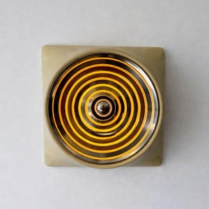 Könnte beinhalten: Eine quadratische, beigefarbene Leuchte mit einem kreisförmigen, konzentrischen Design. Die Mitte des Kreises hat ein kleines, silberfarbenes Element. Die Leuchte strahlt ein warmes, gelbes Licht aus.