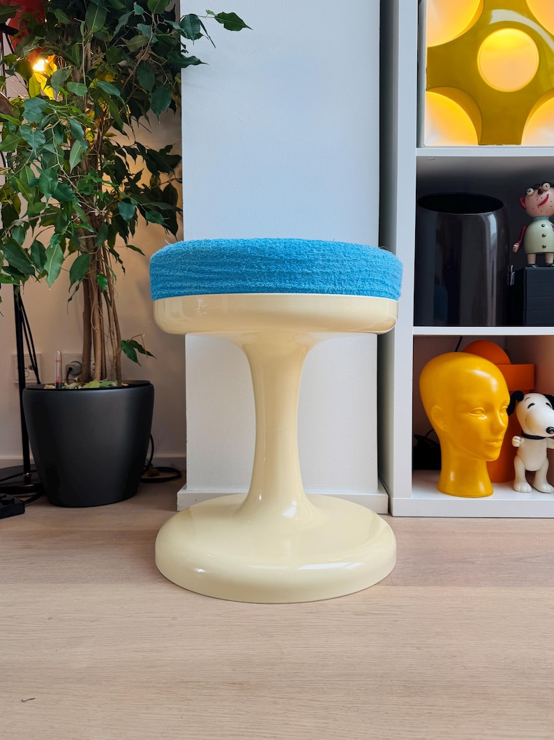 May include: A retro-style stool with a cream-colored base and stem, topped with a round, light blue cushioned seat. The stool has a tulip-like shape and is placed on a wooden floor. The background includes a plant and a shelf with decorative items.