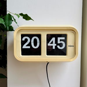 Puede incluir: Un reloj de solapa vintage con marco color crema muestra la hora 20:45. El reloj tiene paneles negros con números blancos y la marca "DEETA" es visible. Un cable de alimentación negro se extiende hacia abajo.