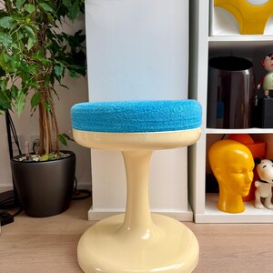May include: A retro-style stool with a cream-colored base and a round, blue, textured seat. The stool has a pedestal-style stem. The background includes a potted plant and a white shelf with decorative items.