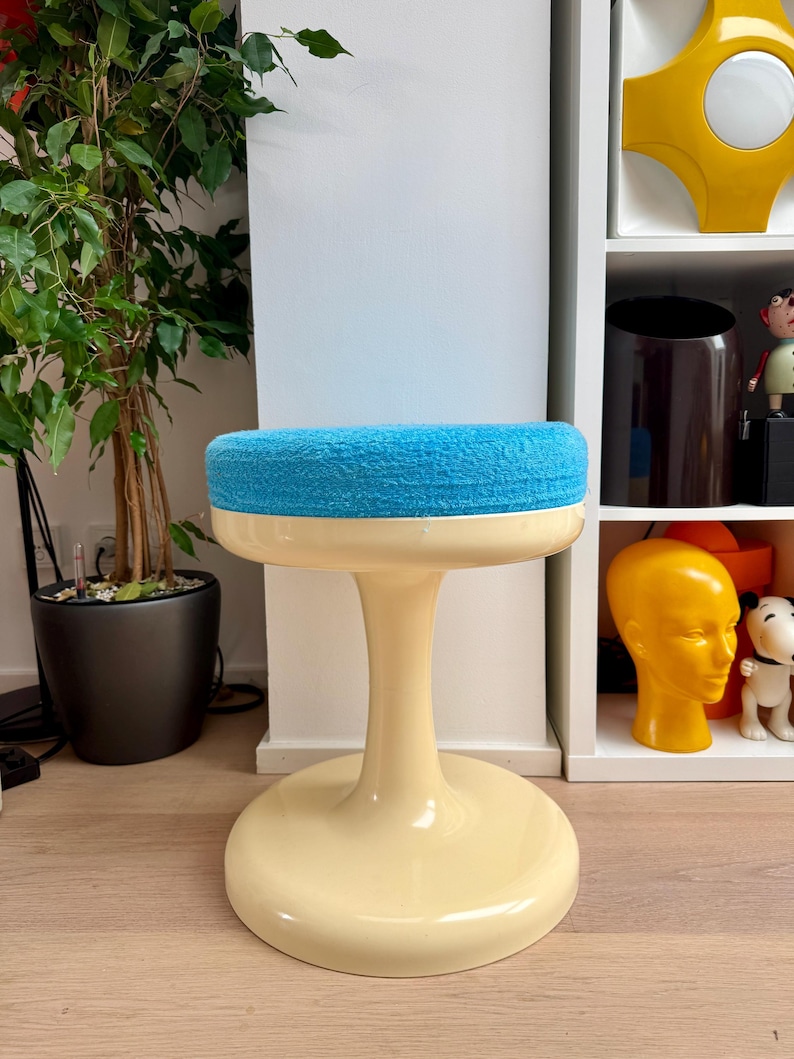 May include: A retro-style stool with a cream-colored base and a round, blue, textured seat. The stool has a pedestal-style stem. The background includes a potted plant and a white shelf with decorative items.