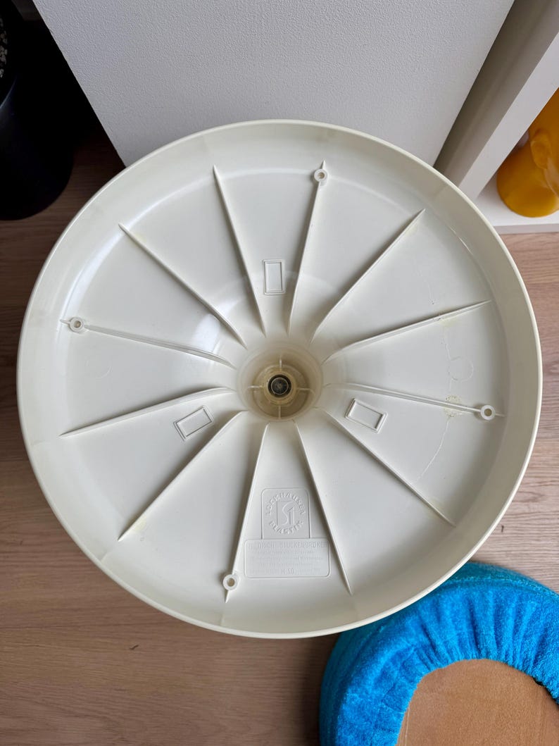 May include: Off-white plastic chair base with a radial spoke design. The base has a central hub with a metal component and several rectangular and circular openings. A blue fabric cushion is visible in the lower right corner.