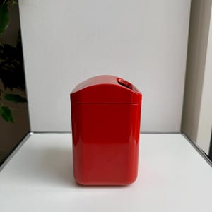 Puede incluir: Un recipiente rectangular rojo brillante con una parte superior curvada y una abertura para el asa. La superficie brillante refleja la luz, resaltando su textura suave. El recipiente es probablemente para almacenamiento o exhibici&oacute;n.