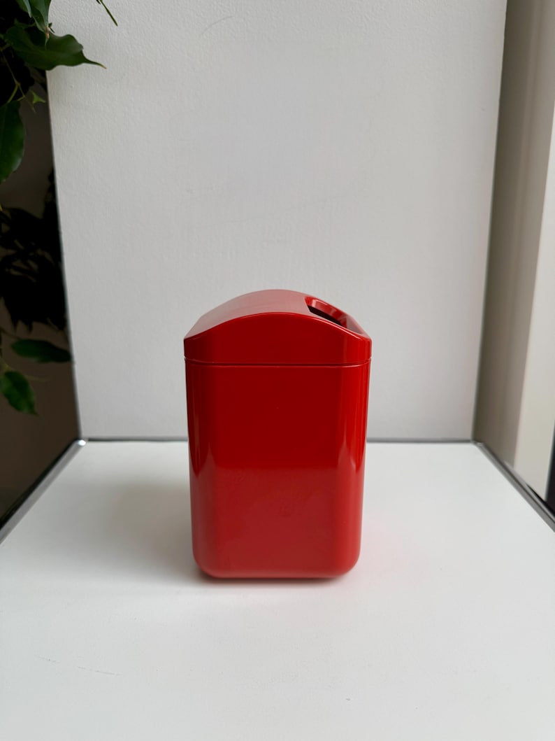 Puede incluir: Un recipiente rectangular rojo brillante con una parte superior curvada y una abertura para el asa. La superficie brillante refleja la luz, resaltando su textura suave. El recipiente es probablemente para almacenamiento o exhibici&oacute;n.