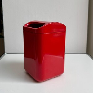 Puede incluir: Un contenedor rectangular rojo brillante con una parte superior curva y un interior negro. El contenedor tiene esquinas redondeadas y una superficie lisa y reflectante. El objeto es un cubo de basura.