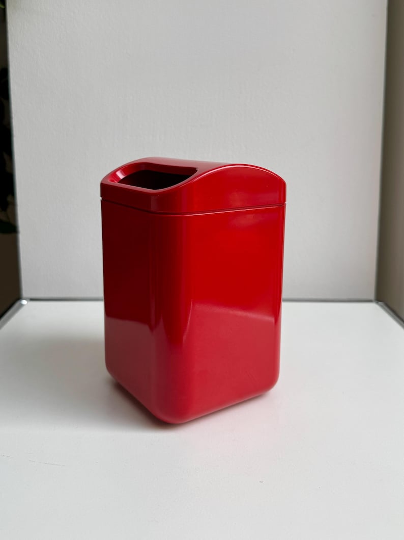 Puede incluir: Un contenedor rectangular rojo brillante con una parte superior curva y un interior negro. El contenedor tiene esquinas redondeadas y una superficie lisa y reflectante. El objeto es un cubo de basura.