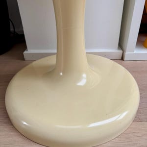 May include: A cream-colored, pedestal-style table base. The base has a round, flat bottom and a curved, column-like stem. The glossy finish reflects light, highlighting the smooth, rounded forms. The table base is a mid-century modern design.