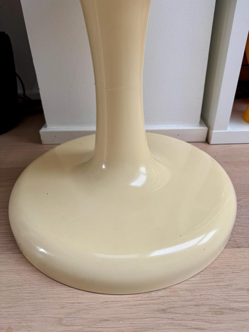 May include: A cream-colored, pedestal-style table base. The base has a round, flat bottom and a curved, column-like stem. The glossy finish reflects light, highlighting the smooth, rounded forms. The table base is a mid-century modern design.