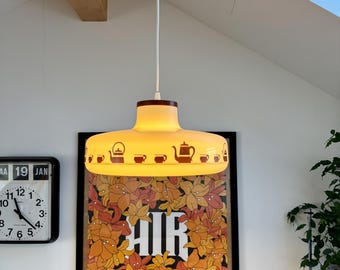 May include: A vintage yellow pendant light with a brown top and a white cord. The light fixture has a design of teapots and cups around the edge. Below the light is a framed movie poster with the text "AIR" and "The Virgin Suicides".