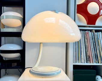 May include: A retro mushroom-shaped table lamp with a cream shade and a curved stem. The base is a light blue circle. The lamp is on a white cube, with other retro lamps on shelves in the background.