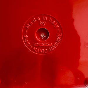 Puede incluir: Primer plano de una superficie roja brillante con el texto en relieve "Made in Italy by gady MAKIO HASUIKE". El texto est&aacute; dispuesto en un patr&oacute;n circular alrededor de un agujero central.