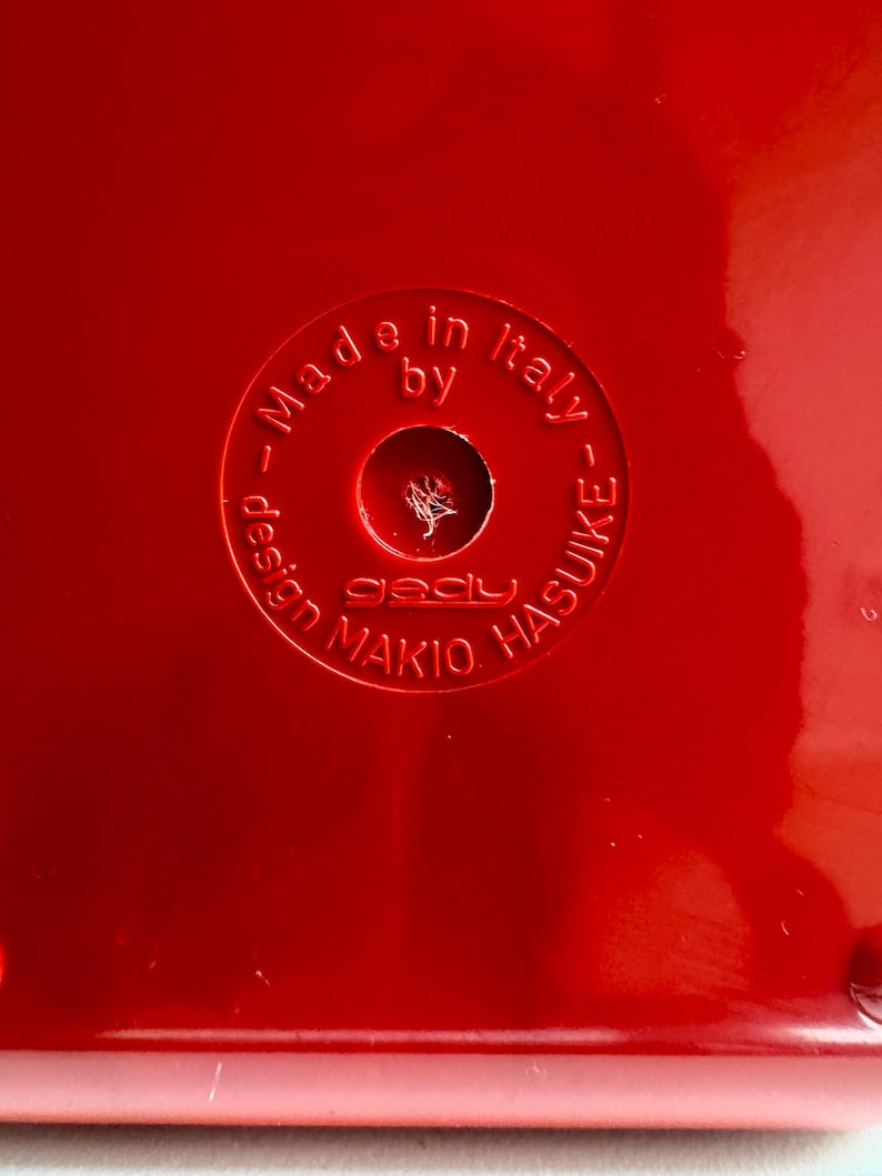 Puede incluir: Primer plano de una superficie roja brillante con el texto en relieve "Made in Italy by gady MAKIO HASUIKE". El texto est&aacute; dispuesto en un patr&oacute;n circular alrededor de un agujero central.