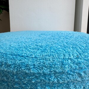 May include: A round, light blue, plush cushion. The cushion has a soft, textured surface and is likely made of a looped fabric. The cushion is a solid color and appears to be new.