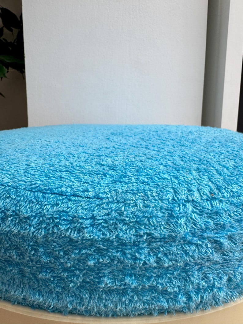 May include: A round, light blue, plush cushion. The cushion has a soft, textured surface and is likely made of a looped fabric. The cushion is a solid color and appears to be new.