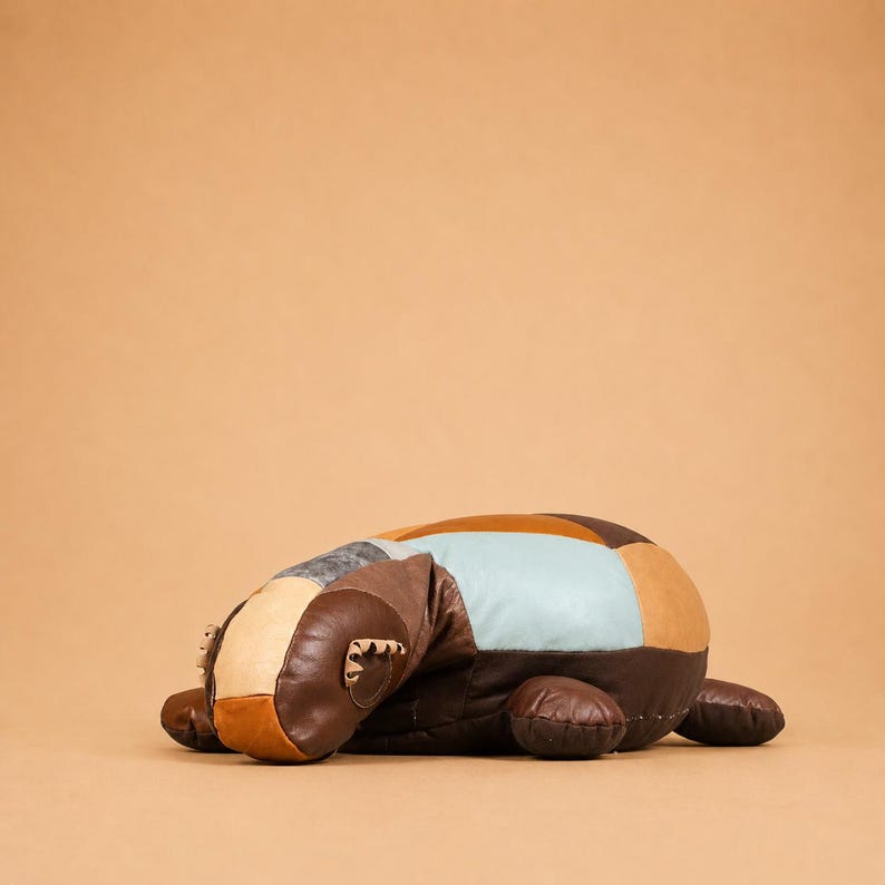 May include: A plush, patchwork badger-shaped pillow in shades of brown, tan, and blue. The pillow has a rounded body and small, rounded legs. The badger's head features small, rounded ears and a detailed snout.