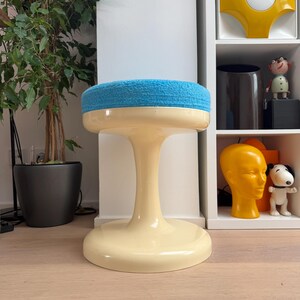 May include: A retro-style stool with a cream-colored base and a round, light blue cushioned seat. The stool has a pedestal-style base and a flared foot. The background includes a houseplant and a white shelf with decorative items.