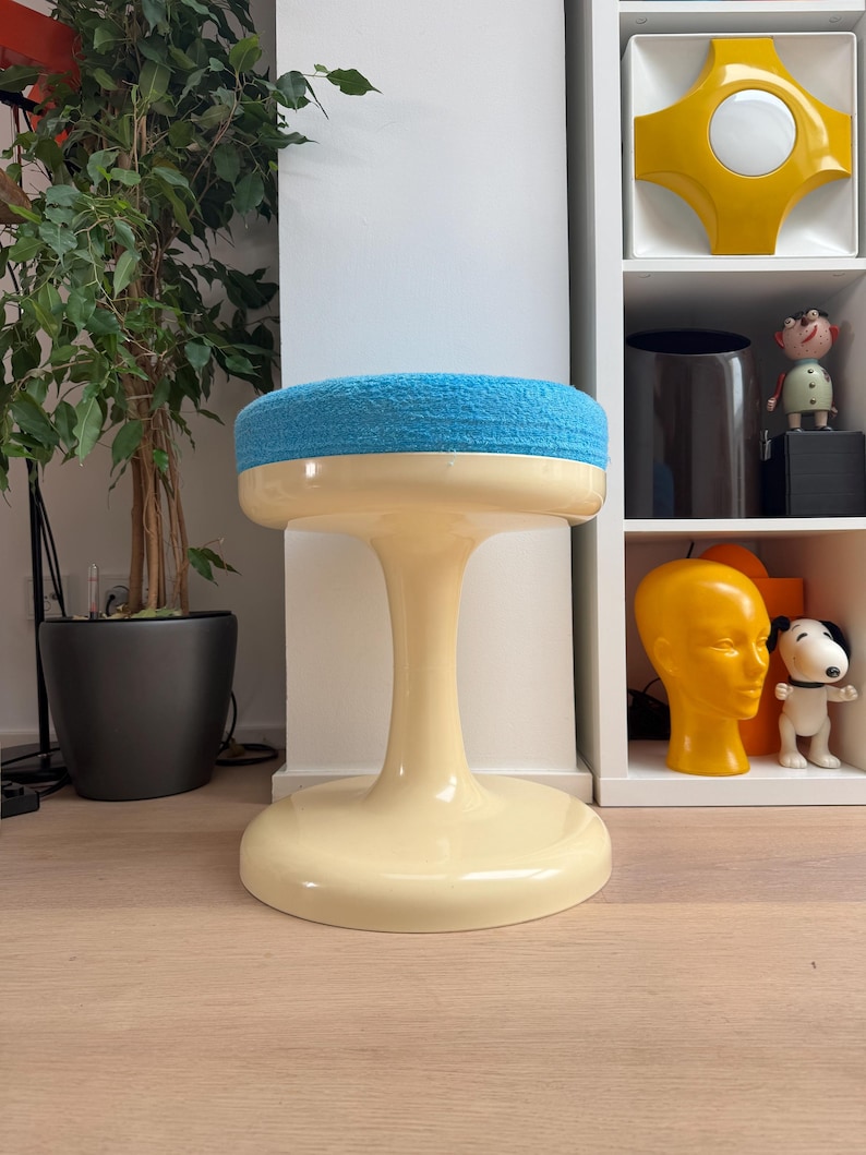 May include: A retro-style stool with a cream-colored base and a round, light blue cushioned seat. The stool has a pedestal-style base and a flared foot. The background includes a houseplant and a white shelf with decorative items.
