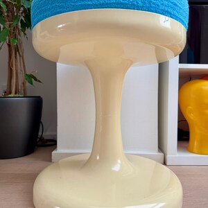 May include: A retro-style stool with a cream-colored base and a vibrant blue, plush seat. The stool has a distinctive, rounded shape, with a wide base and a slender, curved stem. The seat is a soft, textured material.