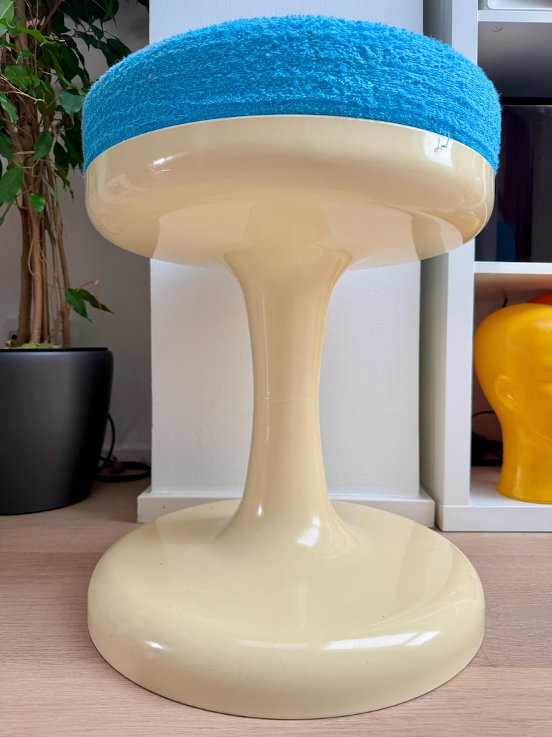 May include: A retro-style stool with a cream-colored base and a vibrant blue, plush seat. The stool has a distinctive, rounded shape, with a wide base and a slender, curved stem. The seat is a soft, textured material.