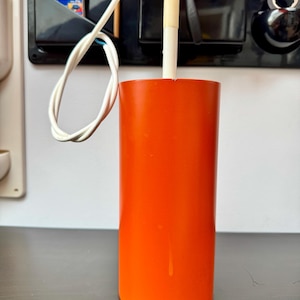 May include: Vibrant orange cylindrical lamp fitting with a white cable and a white cylindrical part. The lamp is on a dark surface, with a black organiser and other items in the background.