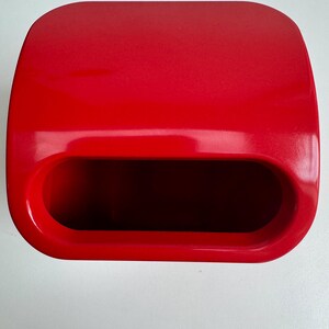 Puede incluir: Un contenedor rectangular rojo brillante con una parte superior redondeada y una gran abertura ovalada. El contenedor tiene un acabado liso y brillante y parece estar hecho de pl&aacute;stico. La abertura revela un interior oscuro.